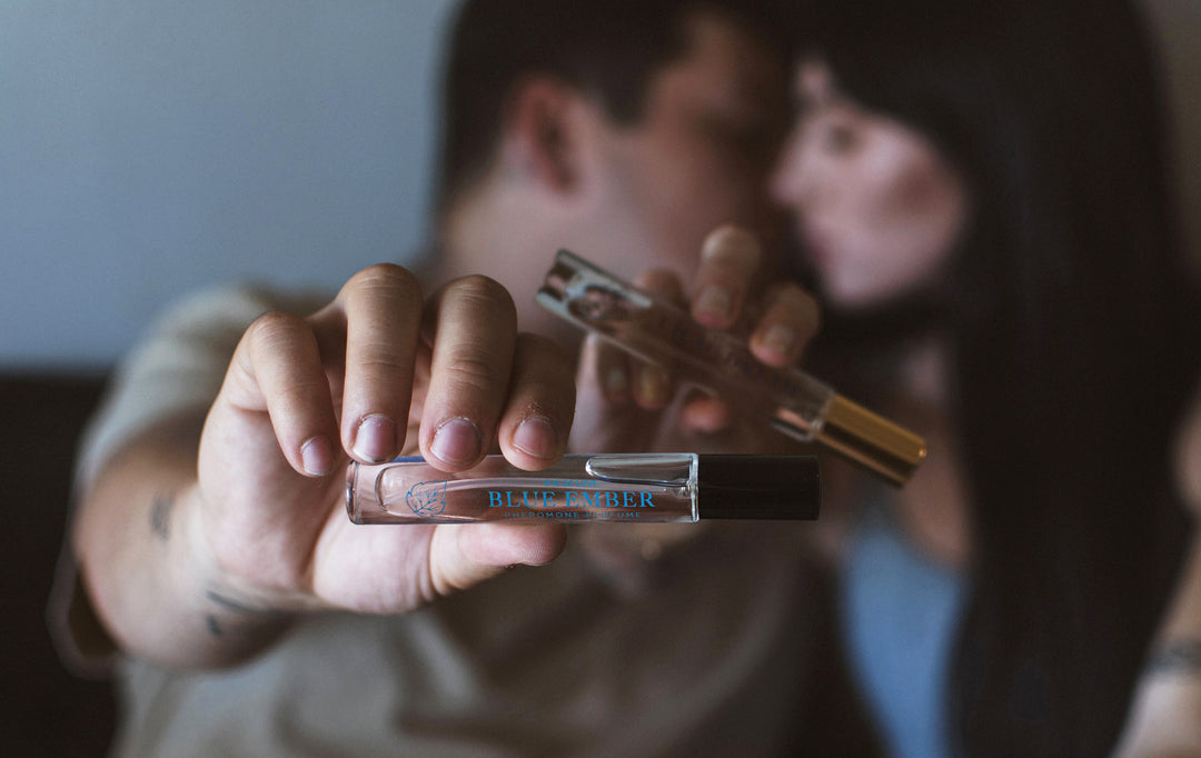 kissing couple in the background holding a bottle of Blue Ember and Lilac Dream pheromone scents to the camera